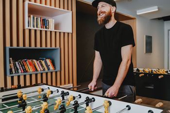 A man with a beard is playing foosball in a room with a bookshelf.